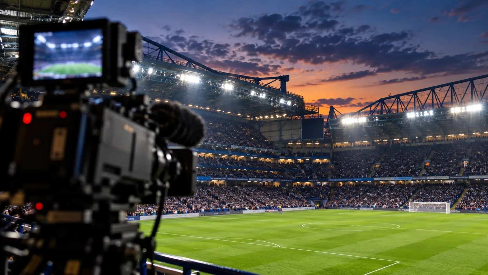 Câmera de transmissão em primeiro plano à esquerda diante de um campo de futebol iluminado e arquibancadas cheias ao entardecer.